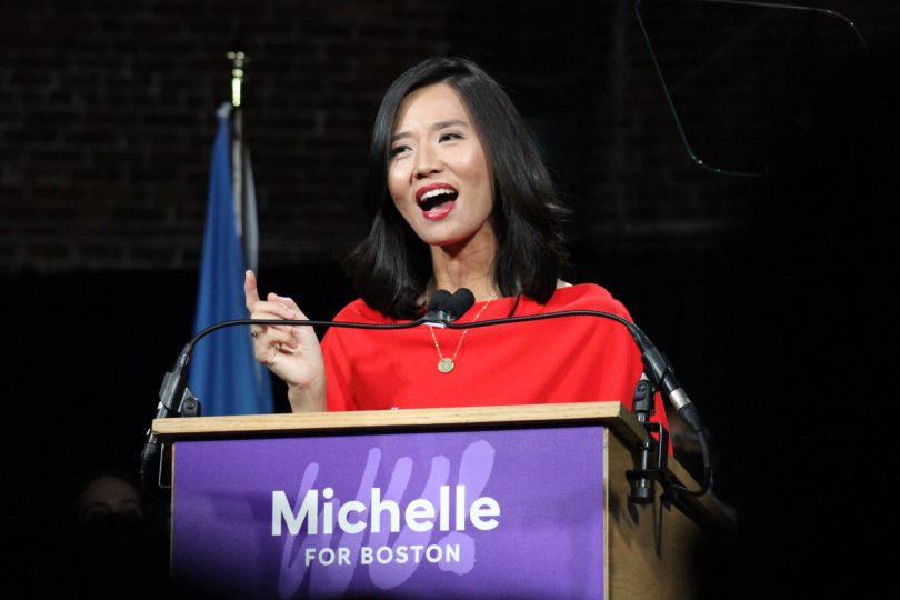 Michelle Wu wearing a red dress stands in front of a podium with a purple sign that reads "Michelle for Boston."