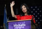 Michelle Wu wearing a red dress stands in front of a podium with a purple sign that reads "Michelle for Boston." She is waving to the audience.