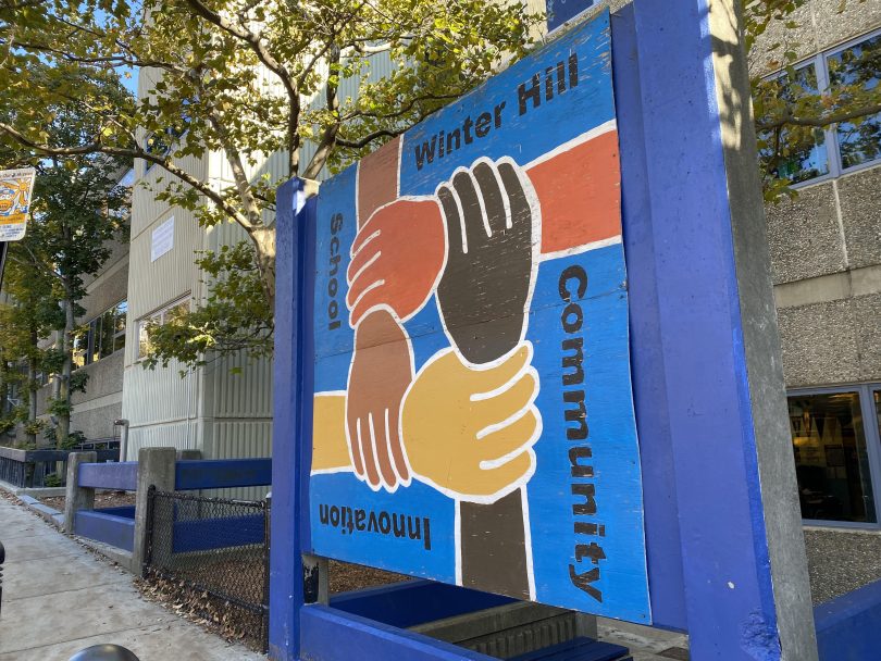 A blue sign showing four hands in shades of orange and brownall holding each others' wrists with the words "winter hill community center"