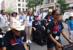 Charlie Carr, a white, older man, is sat in a wheelchair, among volunteers and activists wearing black t shirts with the words ADA 25 in red and blue letters.